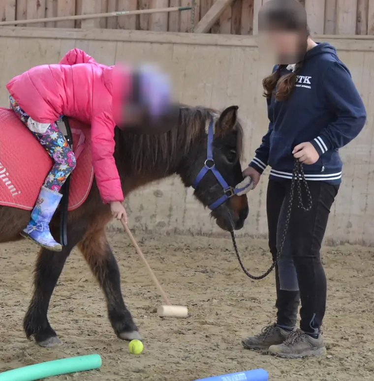 In der Reithalle beugt sich ein Kind vom Pony und spielt mit einem Holzschläger einen Ball, daneben führt eine Betreuerin das Pony