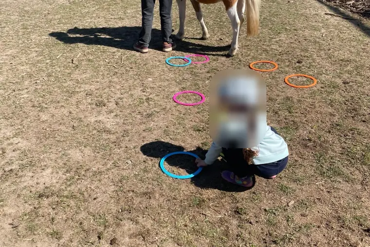Auf einer Wiese liegen bunte Reifen, ein Mädchen mit Helm hockt daneben, dahinter wird ein Pony mit einem Kind im Sattel geführt